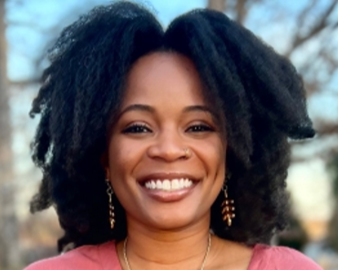 The image shows a smiling Black woman with dark, voluminous hair. She is wearing a pink top and earrings. The background is blurred, suggesting an outdoor setting. Her expression is warm and inviting.